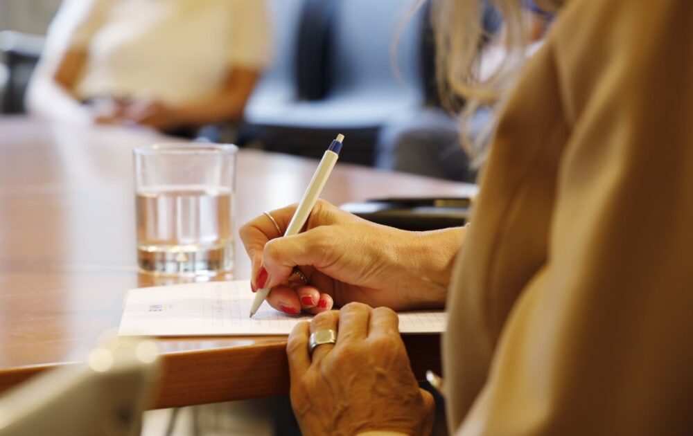 Bild mit zwei einander gegenüber sitzenden Personen. Eine davon ist am Schreiben.